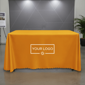 亮橘色平紋布桌巾放置於現代化室內展間，正面展示白色「YOUR LOGO」預留印製區域。具備精緻大花邊剪裁與加厚防皺面料的高質感垂墜細節。
